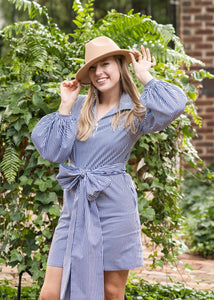 Woman in a blue dress and hat standing outdoors near a brick wall and greenery