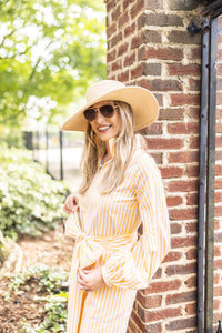 Woman in a yellow dress and hat standing against a brick wall with greenery.