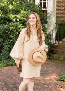 Woman in a beige dress holding a hat on a brick path in front of a brick building.