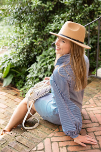Woman sitting on a brick path wearing a beige hat and striped outfit with a handbag.