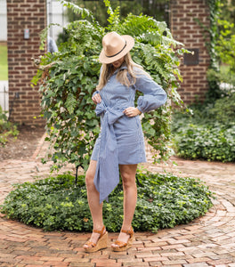 Woman in a blue dress and hat standing outdoors on a brick path with greenery.